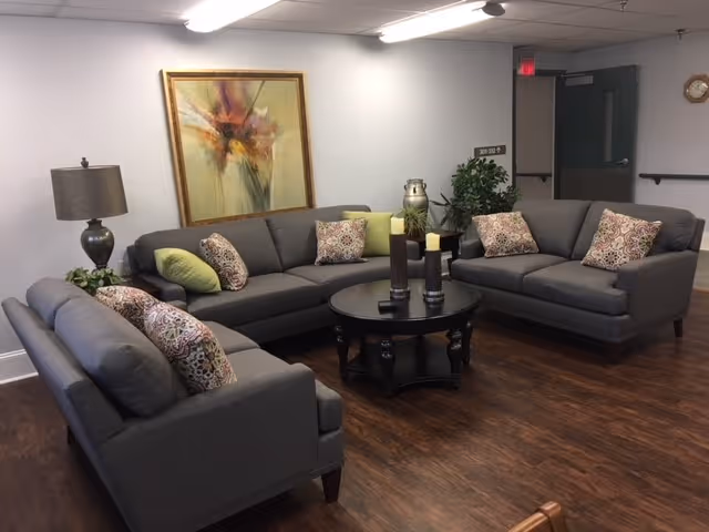 A cozy living room area with three gray sofas arranged around a round black coffee table. The sofas have decorative pillows in green and patterned designs. There is a table lamp with a dark base and a beige shade on a side table, a large abstract painting on the wall, and some plants in the corner. The floor is wooden, and the walls are light-colored. A clock is mounted on the wall near a door in the background.