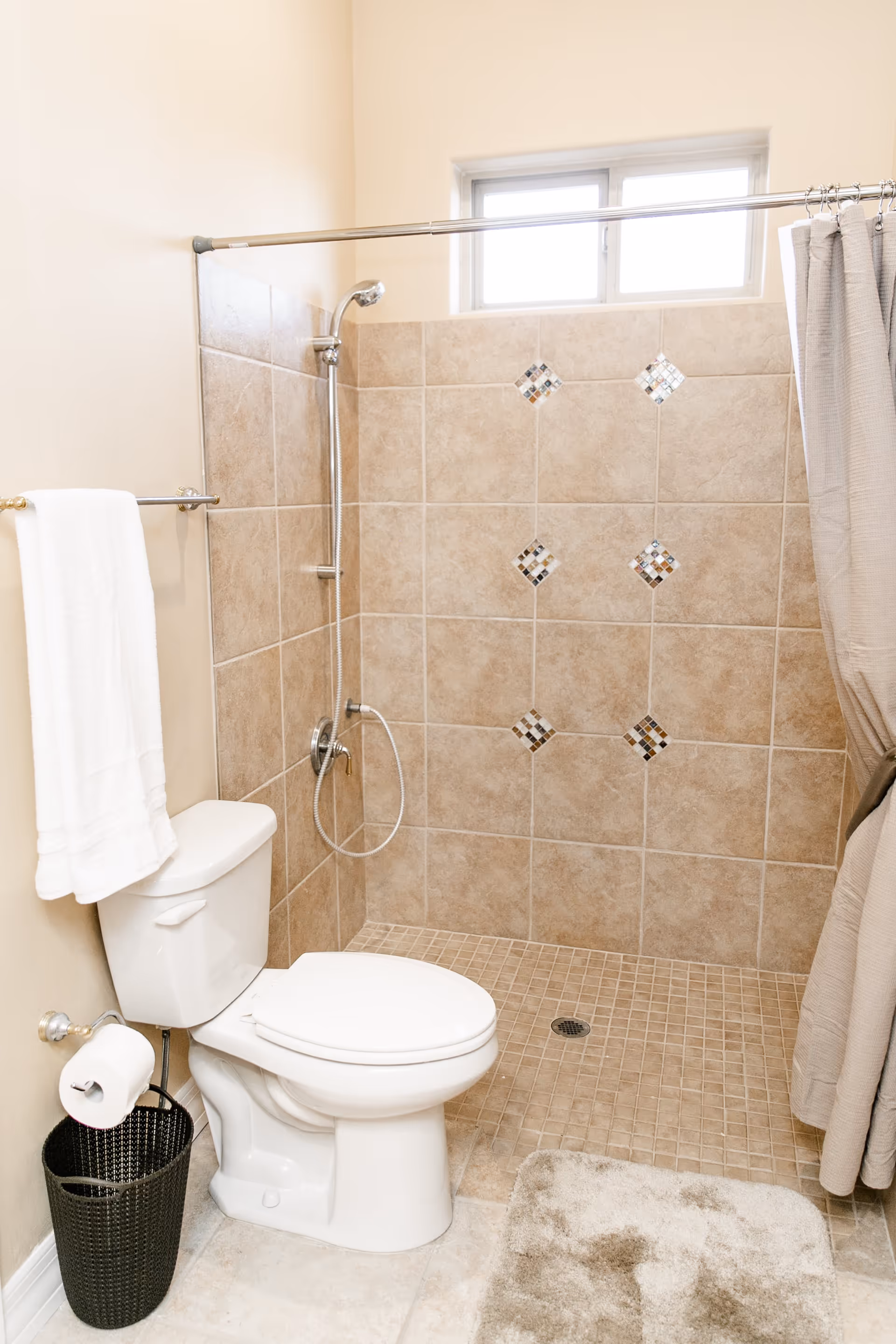 Tiled bathroom with a toilet, towel, waste basket and a walk-in shower with a handheld showerhead and curtain.