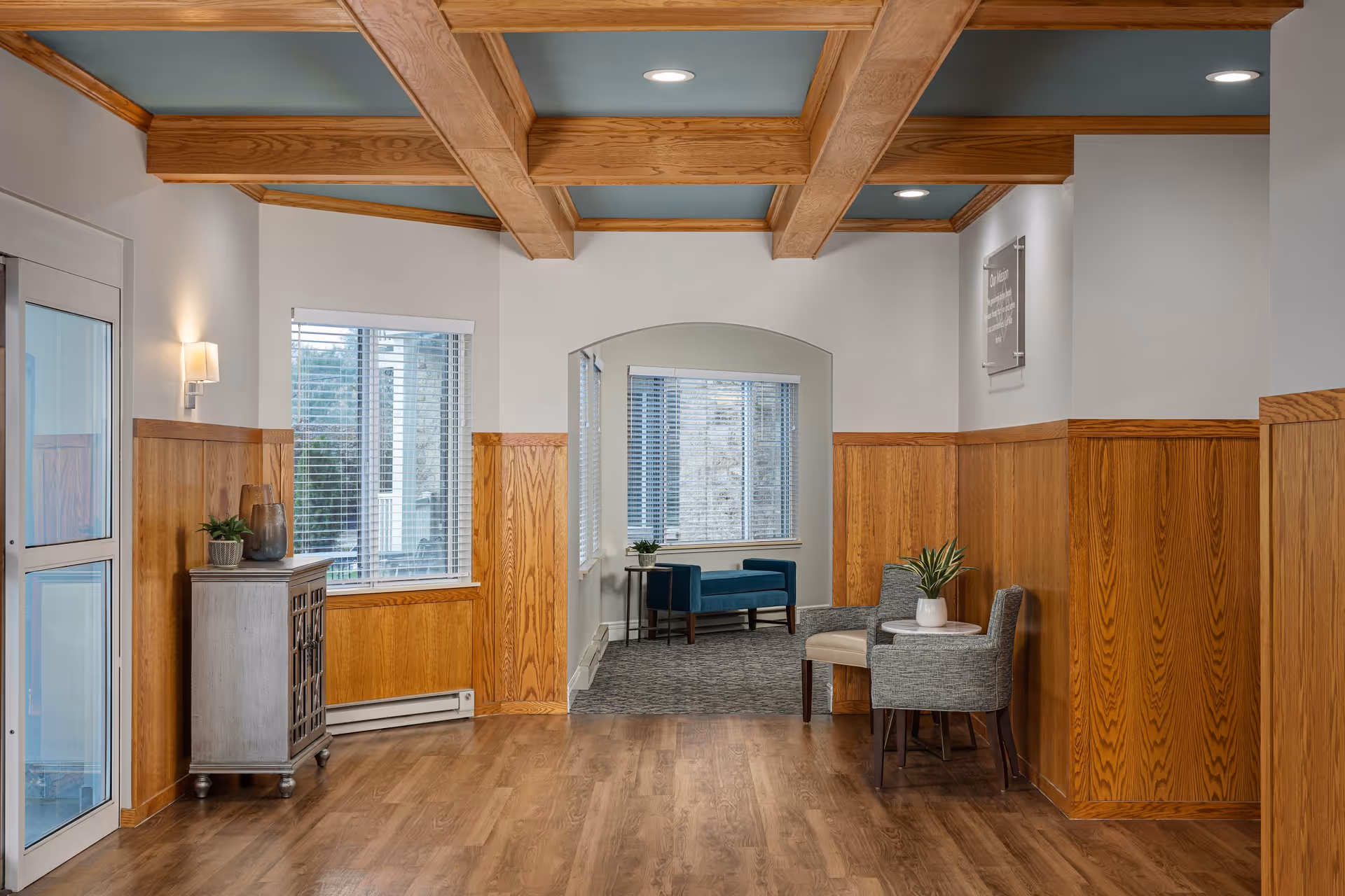 Interior view of a senior living facility hallway with wooden paneling on the walls and wooden beams on the ceiling. There is a small cabinet with decorative items on the left, a seating area with two chairs and a small table with a plant on the right, and a blue bench near a window in the background.