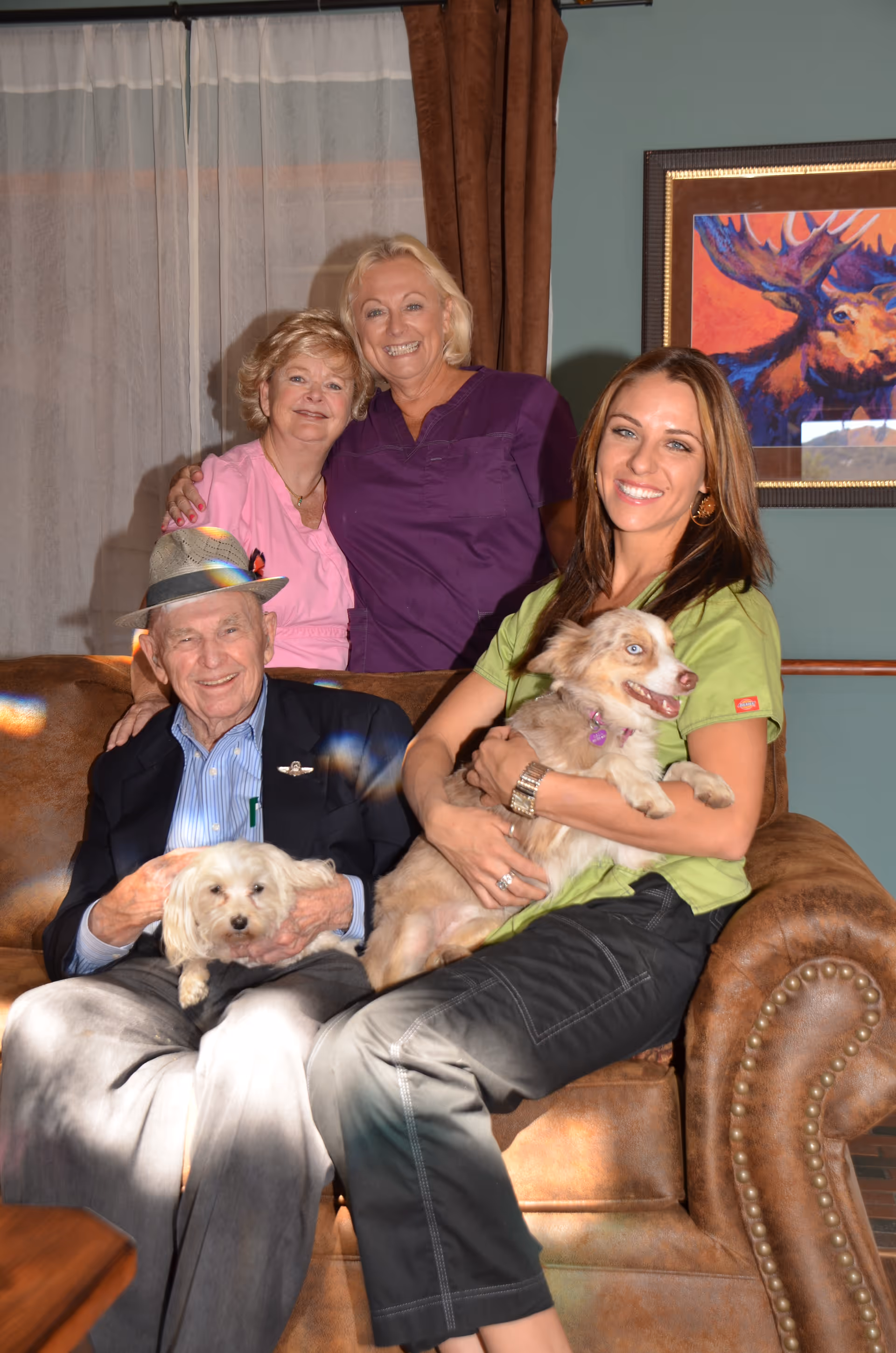 Four people sitting and standing in a cozy living room setting. An elderly man wearing a hat and a dark blazer is seated on a brown leather couch holding a small white dog. A younger woman in a green shirt is sitting next to him on the couch holding a medium-sized dog with light brown and white fur. Behind the couch, two women are standing close together, smiling, one wearing a pink top and the other in a purple top. A colorful framed painting of a moose is visible on the wall behind them.