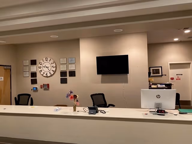 Reception desk area with chairs, a computer, a vase of flowers, a wall clock and a mounted TV.