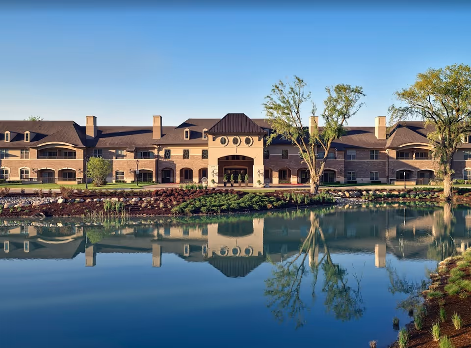 Exterior view of a senior living facility named Flourish Collection at Rochester, showing a large two-story building with a peaked roof and multiple windows. The building is reflected in a calm pond in the foreground, with landscaped greenery and trees around the water. The sky is clear and blue.