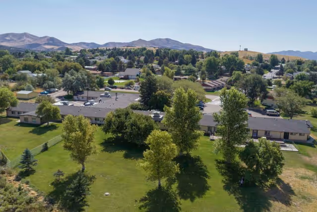 Aerial view of Monte Vista Hills Health Care Center surrounded by green lawns and trees, with mountains in the background under a clear sky.
