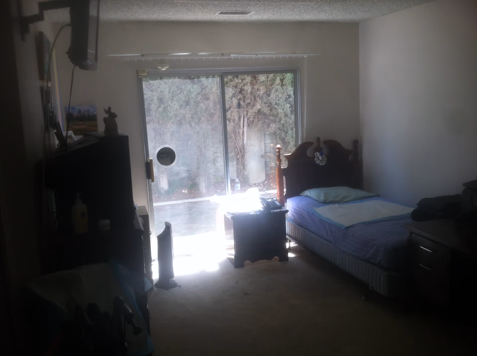 A small bedroom with a single bed against the right wall, a wooden headboard, and a blue bedspread. There is a nightstand next to the bed with a telephone on it. A sliding glass door at the back of the room lets in natural light and shows a view of trees outside. On the left side, there is a TV mounted on the wall above a dresser with various items on it. The room has beige carpet and plain white walls.