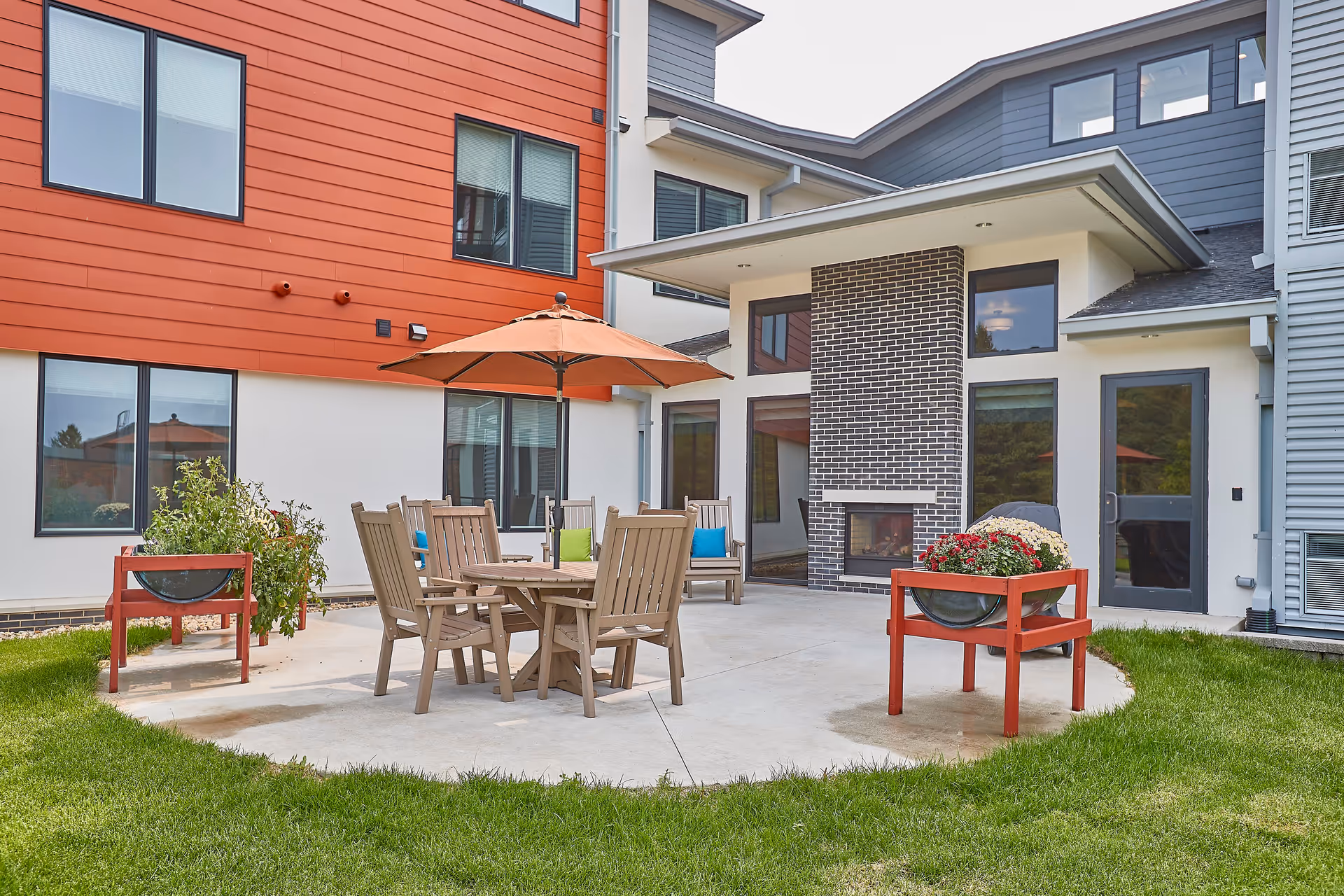 Outdoor patio area at a senior living facility with a round table, several wooden chairs, and an orange umbrella. The patio is surrounded by green grass and features two red planters with flowers. The building exterior has a mix of red, white, and gray siding with large windows and a brick fireplace.