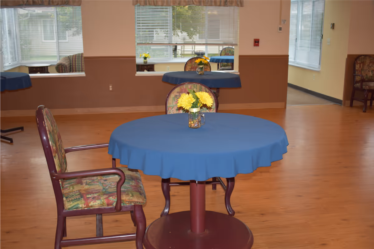 Round table with a blue tablecloth and a vase of yellow flowers in a communal dining area with chairs and wood flooring.