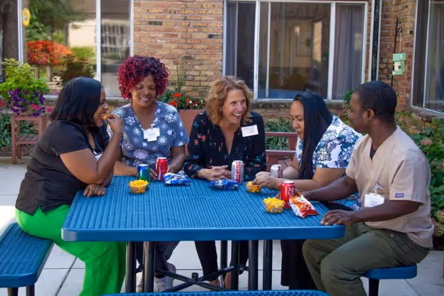 Five people sitting around a blue outdoor picnic table enjoying snacks and drinks, smiling and engaging in conversation. The setting appears to be a courtyard or patio area with brick walls and windows in the background.