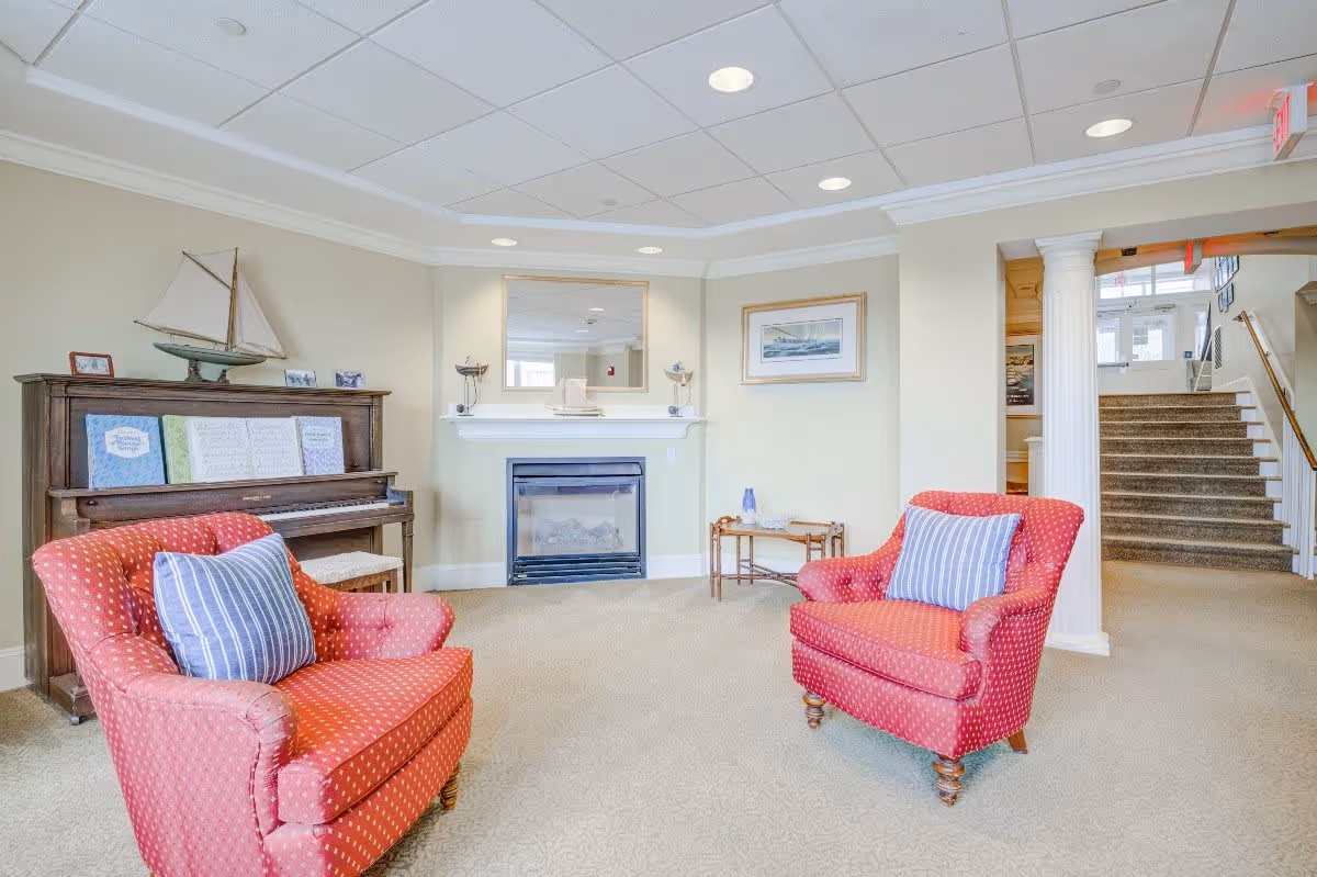 A cozy living room area with two red armchairs featuring blue and white striped pillows, a wooden piano with sheet music and a model sailboat on top, a fireplace with a mirror above it, a framed picture on the wall, and a small table with decorative items. There is a staircase and a hallway visible in the background.