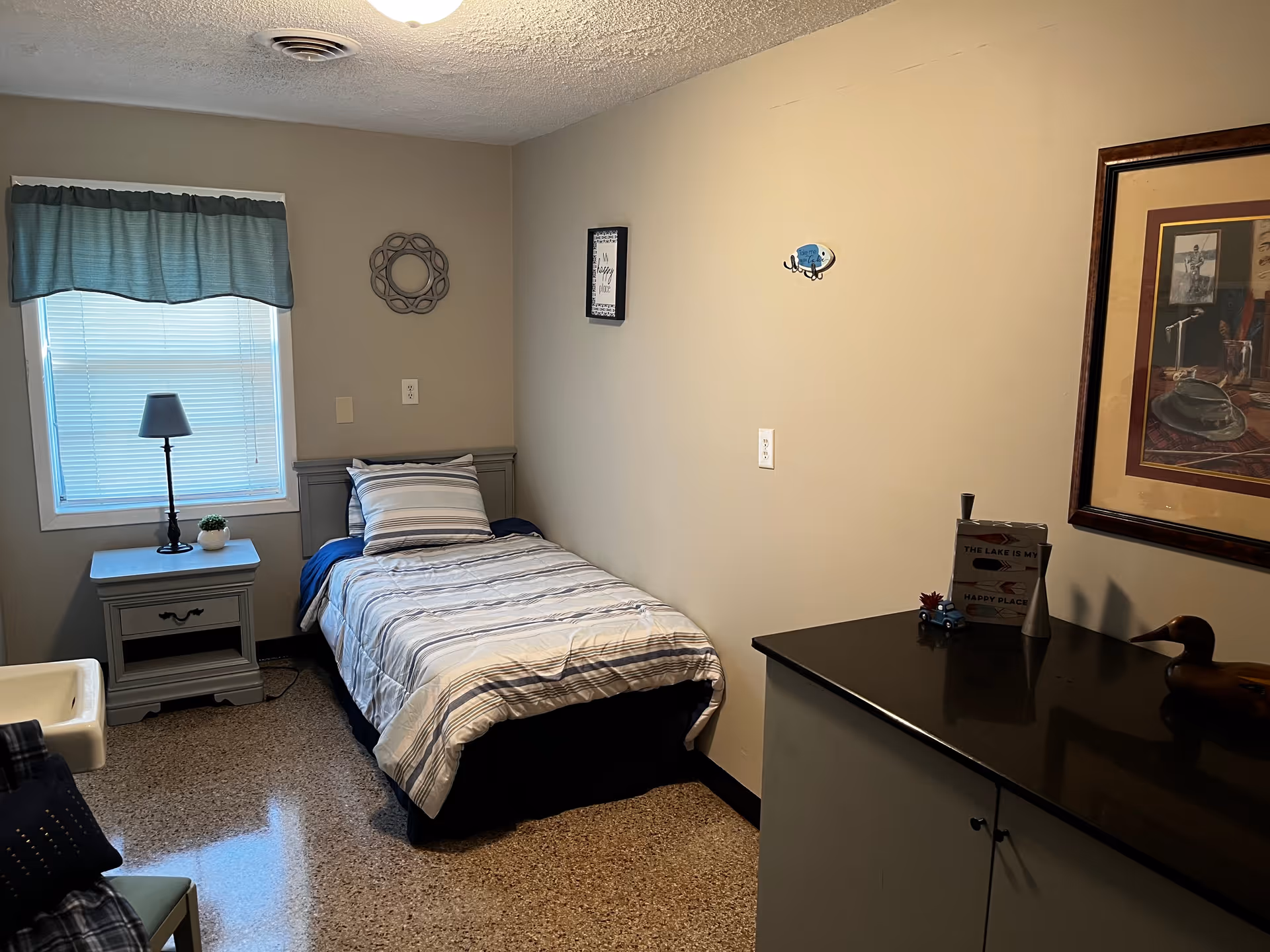 A small, neatly arranged bedroom with a single bed covered in striped bedding. Next to the bed is a nightstand with a lamp and a small plant. A window with a blue valance lets in natural light. On the opposite wall, there is a framed picture and a decorative wall piece. A cabinet with a dark countertop holds a few decorative items, including a small sign and a wooden duck figurine.