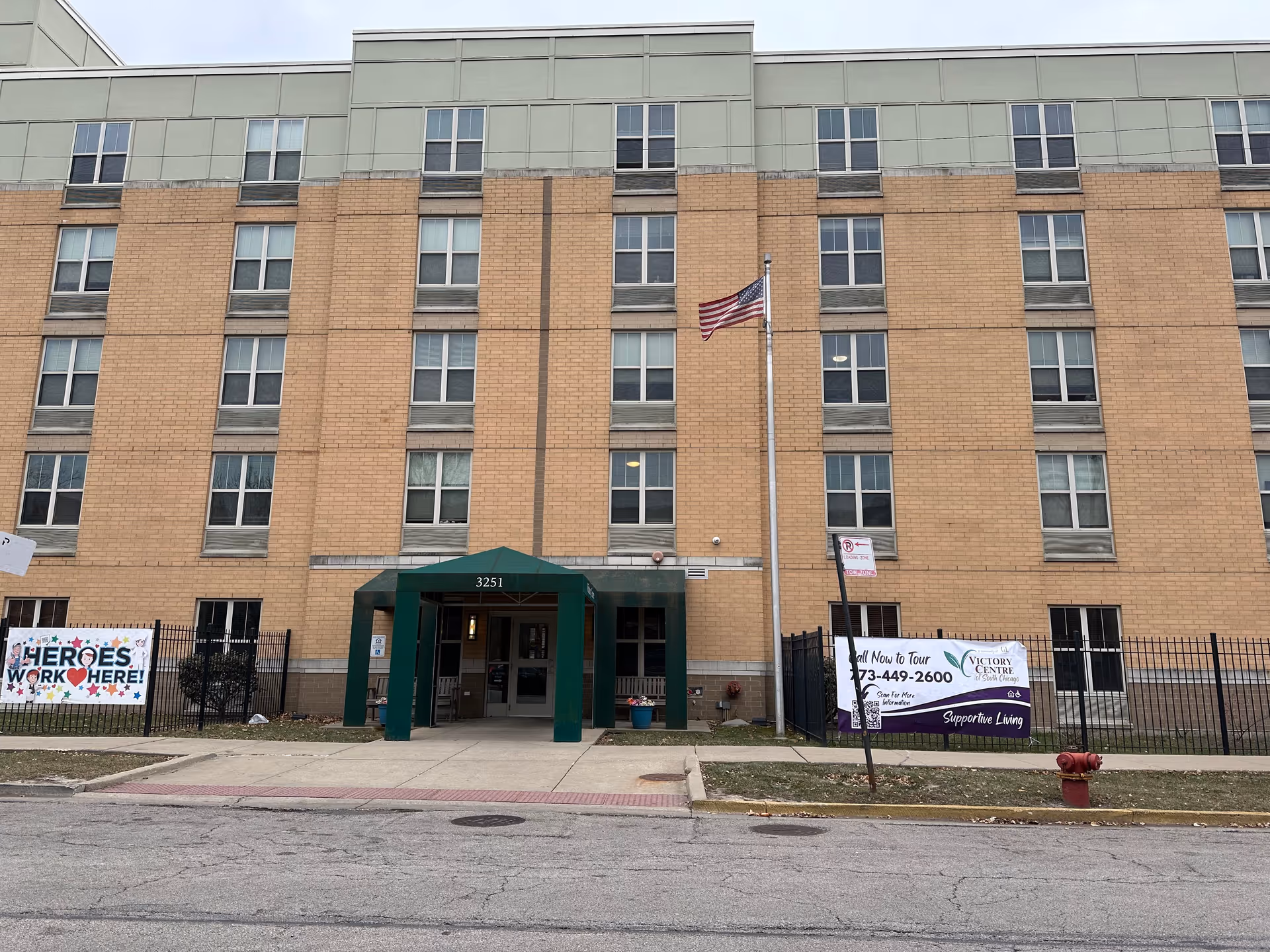Front exterior of a multi-story brick senior living building with a green entrance awning, an American flag, and banners/signs by the sidewalk.
