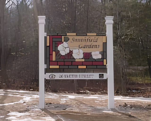 A decorative outdoor sign for Smithfield Gardens Assisted Living featuring a stained glass design with flowers, mounted on two white posts along a snowy path with trees in the background.