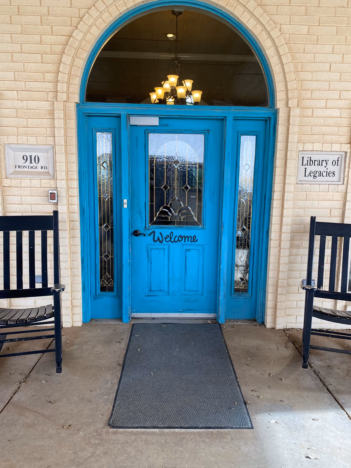 Bright turquoise arched entrance door with decorative glass and a 'Welcome' sign, flanked by rocking chairs and a 'Library of Legacies' plaque.