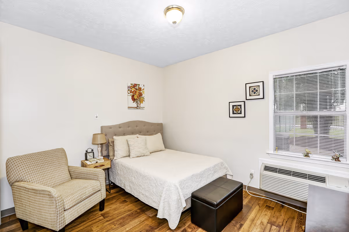 A cozy bedroom with a beige upholstered bed with white bedding and pillows, a beige armchair, a wooden nightstand with a lamp and clock, a window with blinds, and framed artwork on the walls. The room has wooden flooring and a ceiling light fixture.