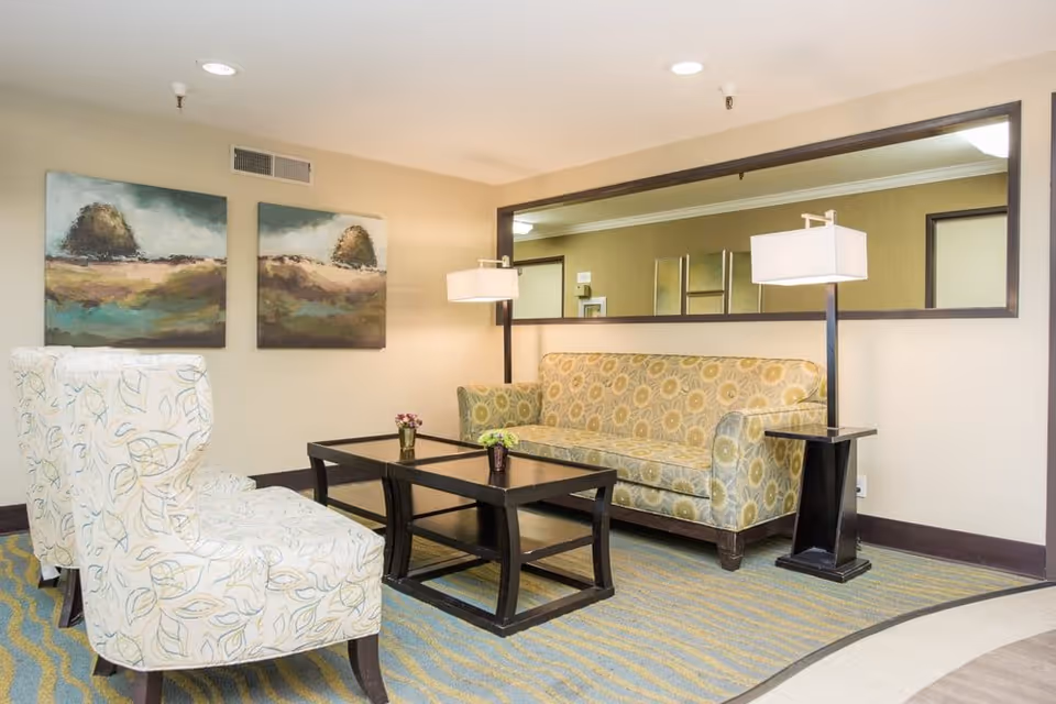 A bright lounge with a patterned sofa and armchairs arranged around a coffee table, wall art, and a large mirror.