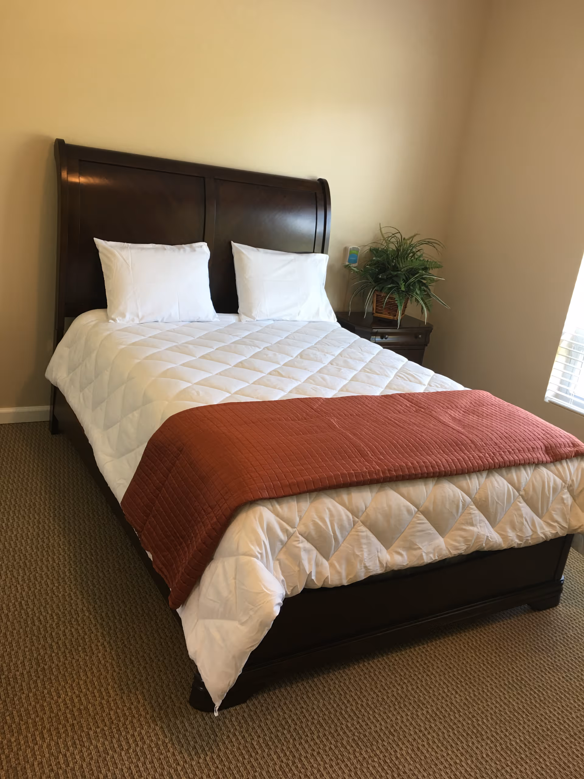 A neatly made bed with a dark wooden headboard, white quilted bedding, two white pillows, and a rust-colored throw blanket at the foot of the bed. There is a small wooden nightstand next to the bed with a green potted plant and a small device on it. The room has beige walls and carpeted floor, with a window letting in natural light.