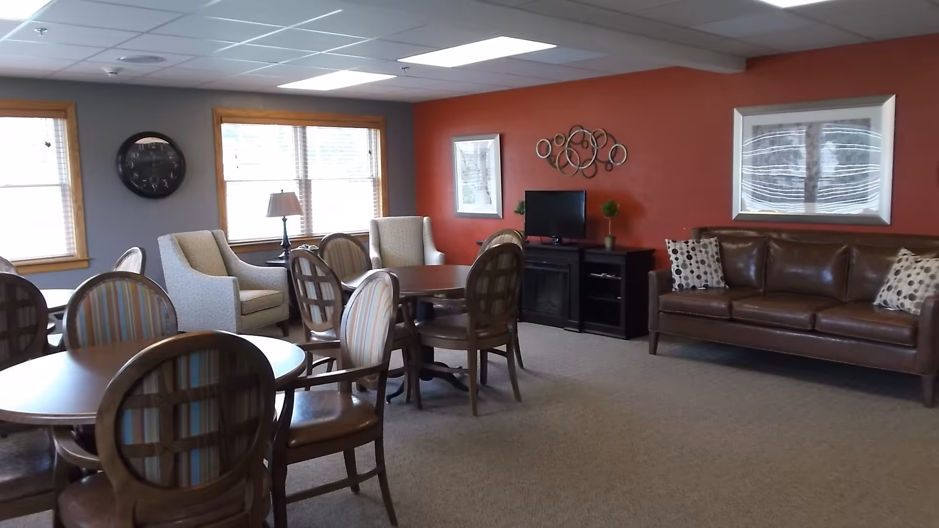 A common area in a senior living facility featuring multiple round tables with wooden chairs, two beige armchairs near windows with wooden frames, a brown leather sofa with patterned pillows against a red accent wall, a small TV on a black stand, and decorative wall art.