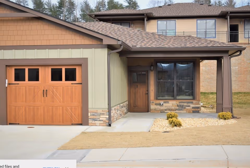 Exterior view of a residential-style building entrance with a wooden garage door, a front door, and a window with blinds. The building features a combination of green siding, stone accents, and brown trim. There is a small landscaped area with rocks and shrubs near the entrance.