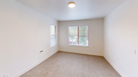 Empty room with beige carpet, white walls, and two windows with blinds letting in natural light.