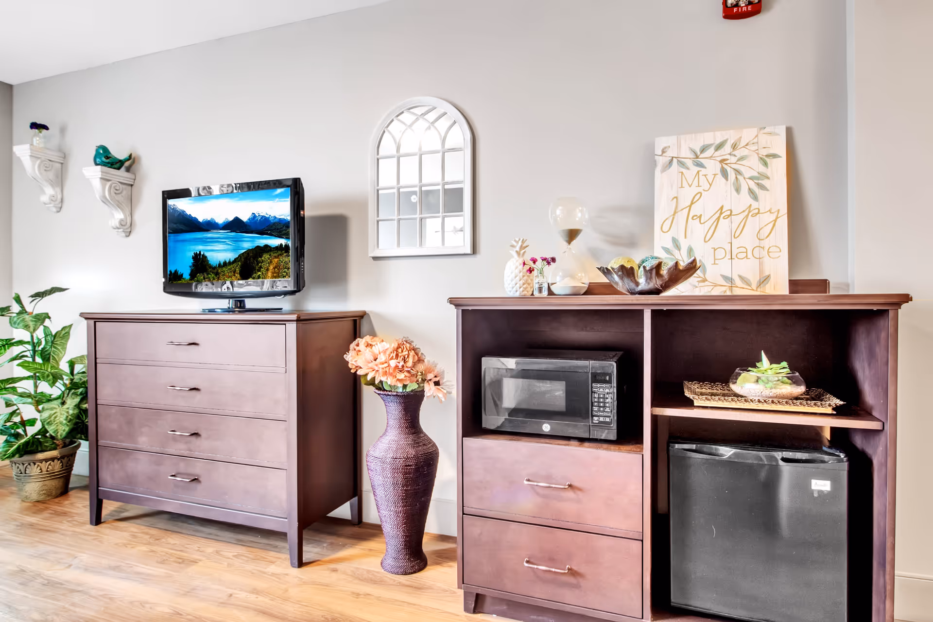 A cozy interior space featuring two wooden cabinets. One cabinet holds a flat-screen TV displaying a scenic mountain and lake view. The other cabinet contains a microwave and a small refrigerator, with decorative items including a vase with flowers, an hourglass, and a sign that reads 'My Happy Place'. There are also wall shelves with small decorative birds, a potted plant, and a wall mirror shaped like a window.
