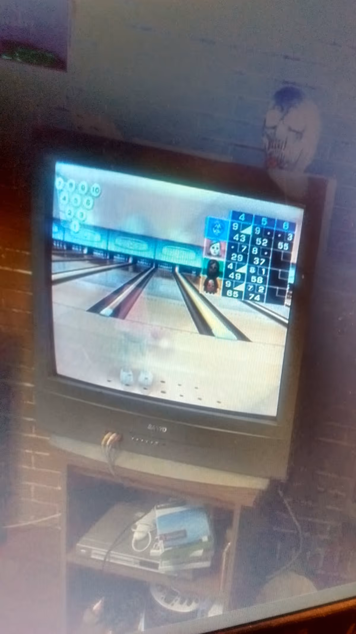 A CRT television displaying a bowling video game screen with scores visible. The TV is placed on a wooden stand with a gaming console and some game cases underneath. The background includes a brick wall and a decorative skull on a shelf.