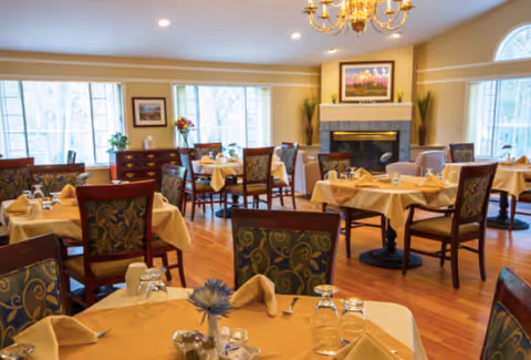 Bright dining room with round tables set with tablecloths and napkins, upholstered chairs, hardwood floors, large windows, and a fireplace.