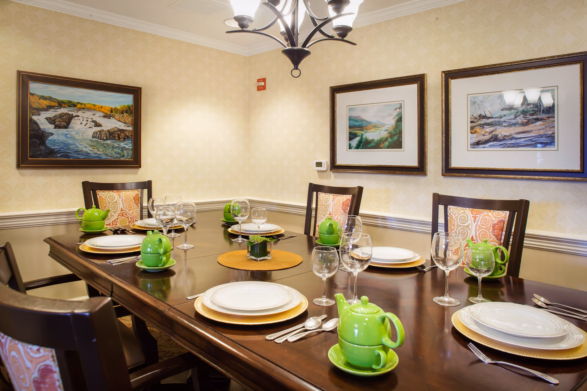 A dining room with a dark wooden table set for six people. Each place setting includes white plates on gold chargers, silverware, wine glasses, and a green teapot with a matching cup. The chairs have patterned cushions with orange and beige designs. The walls are decorated with light yellow patterned wallpaper and three framed landscape paintings. A chandelier with five lights hangs above the table.