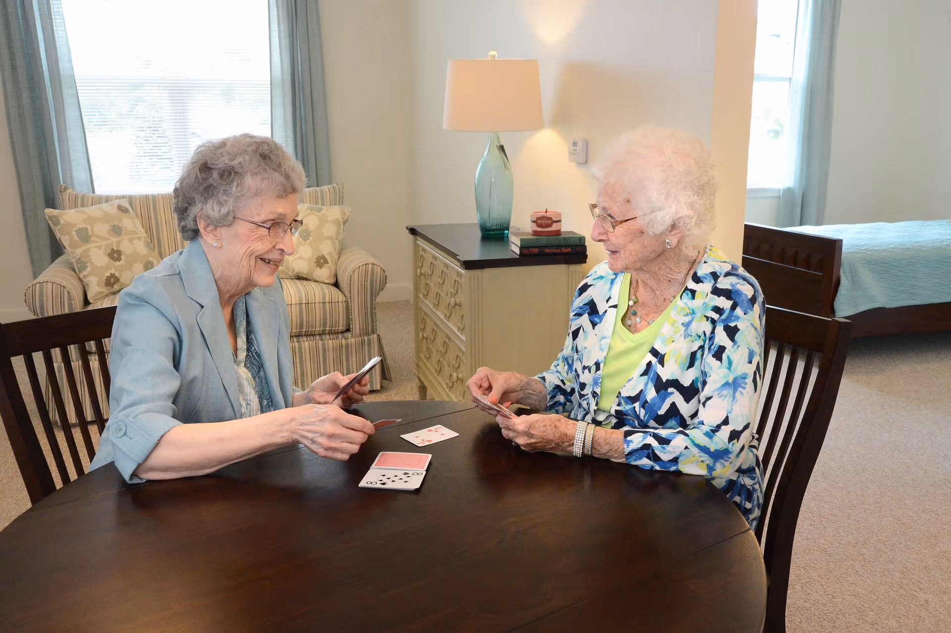 Two elderly women sitting at a round wooden table playing cards in a well-lit room with a bed, armchair, and a lamp on a dresser in the background.
