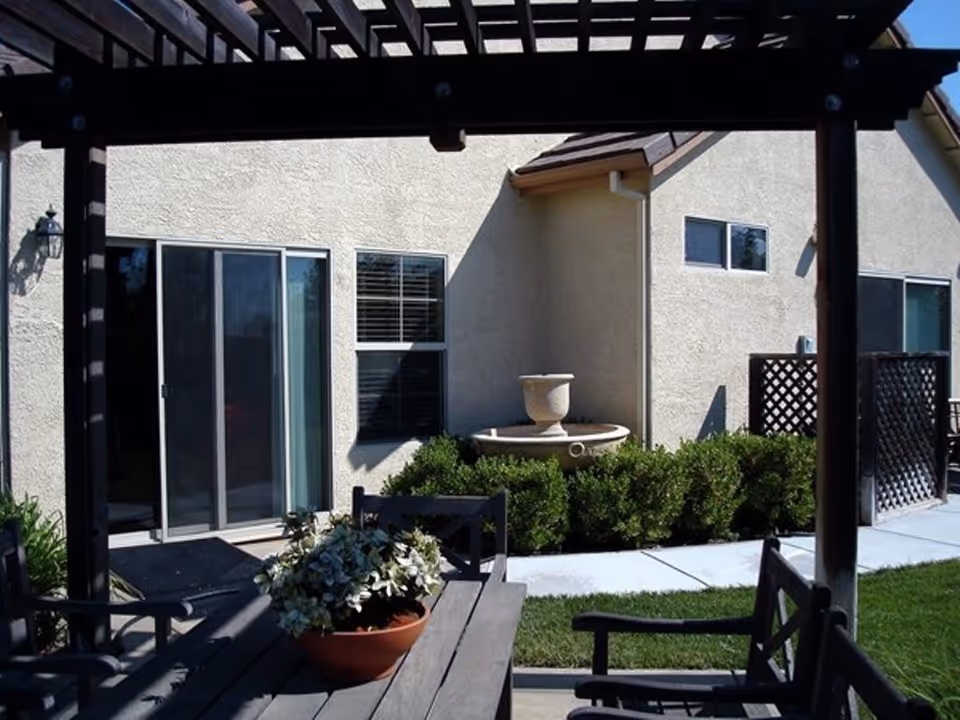 Outdoor patio area with a wooden pergola overhead, a wooden table and chairs, a potted plant on the table, a small fountain surrounded by bushes, and a beige building with sliding glass doors and windows in the background.