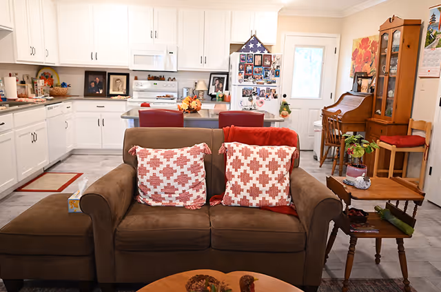 A cozy living room area with a brown sofa featuring two red and white patterned pillows and a red throw blanket. Behind the sofa is a kitchen with white cabinets, a white stove, and a refrigerator covered with photos and magnets. To the right, there is a wooden desk with a chair, a wooden cabinet with glass doors, and a small table with a plant and decorative items. The room has a warm and homey atmosphere.
