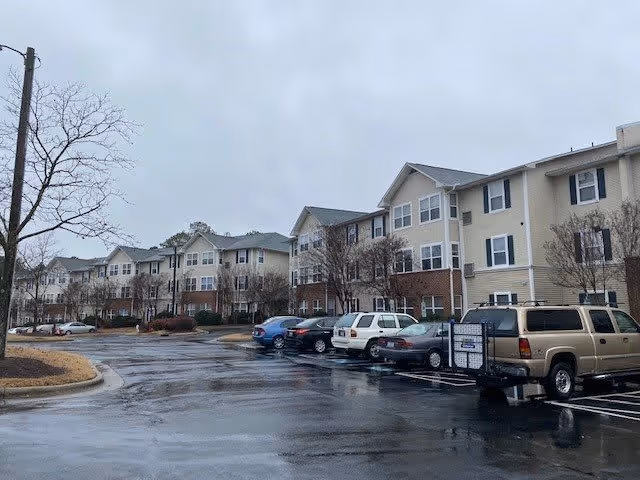 Exterior view of a multi-story senior living facility named Lakeside Gardens on a cloudy day with a wet parking lot in front. Several cars are parked along the building, and leafless trees are visible around the area.