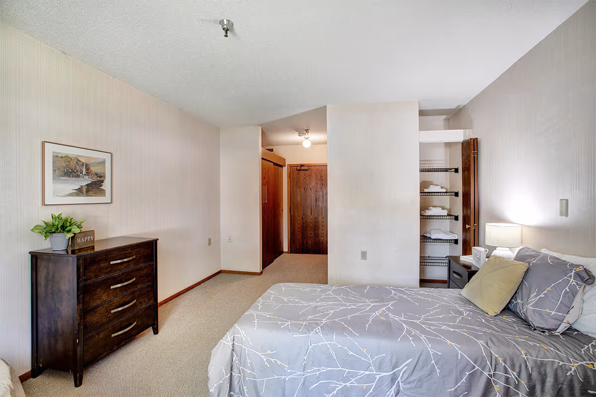A bedroom in a senior living facility featuring a bed with gray and white patterned bedding and pillows, a wooden nightstand with a lamp and a cup, an open closet with shelves holding folded towels, a dark wooden dresser with a small plant and a decorative block that says 'HAPPY', beige carpet, and light-colored walls with a framed picture.