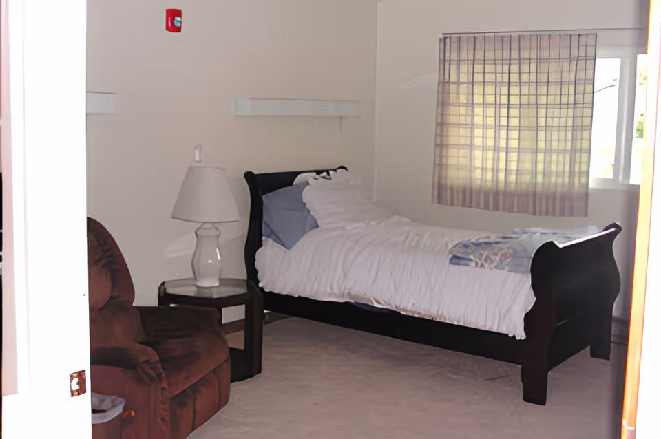 A bedroom with a dark wooden single bed dressed in white linens, a bedside table with a lamp, a brown recliner, and a curtained window.