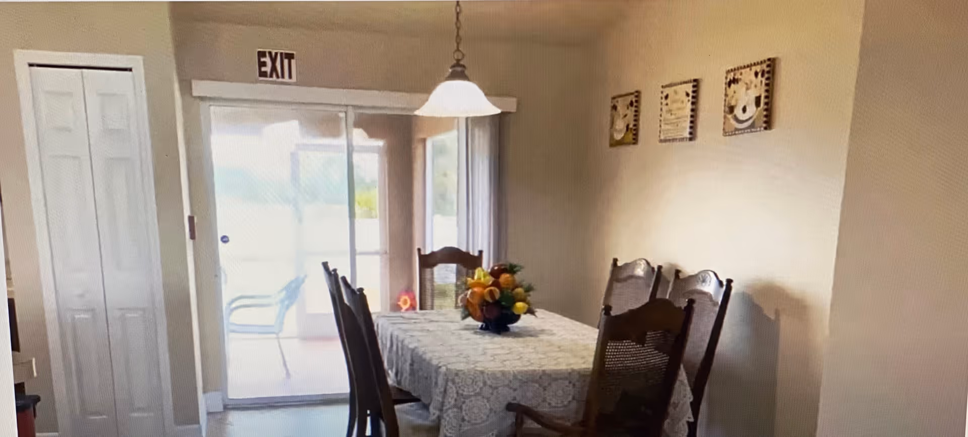 Dining room with a lace-covered table and fruit centerpiece, wooden chairs, a sliding glass door under an EXIT sign, and wall art on a pale wall.