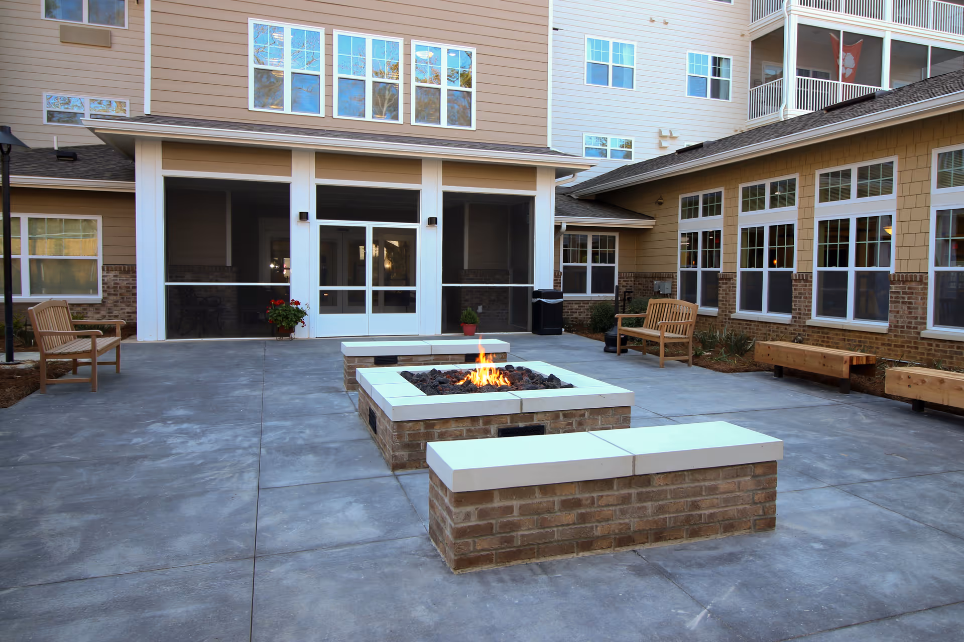 Outdoor courtyard area of Mulligan Park Gracious Retirement Living featuring a central fire pit with a brick base and white stone top, surrounded by wooden benches and chairs. The courtyard is paved with concrete and is adjacent to a multi-story building with large windows and screened doors.