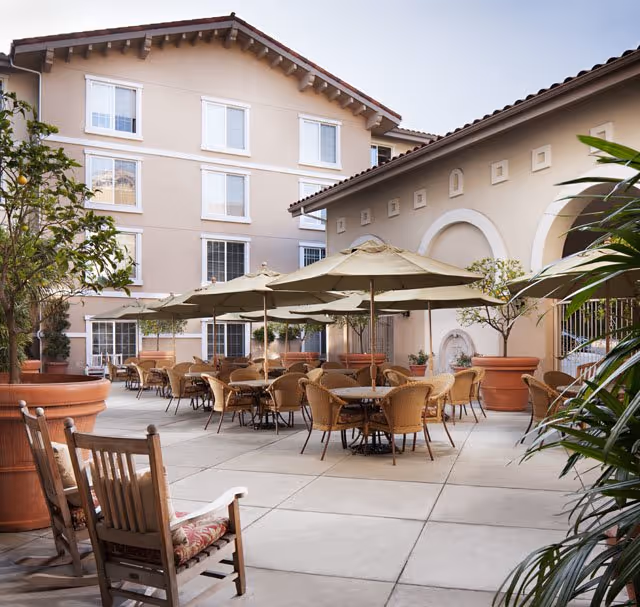 Outdoor patio area at Belmont Village Senior Living Hollywood Hills with multiple tables and chairs under large beige umbrellas, surrounded by potted plants and trees, adjacent to a beige building with windows and architectural details.