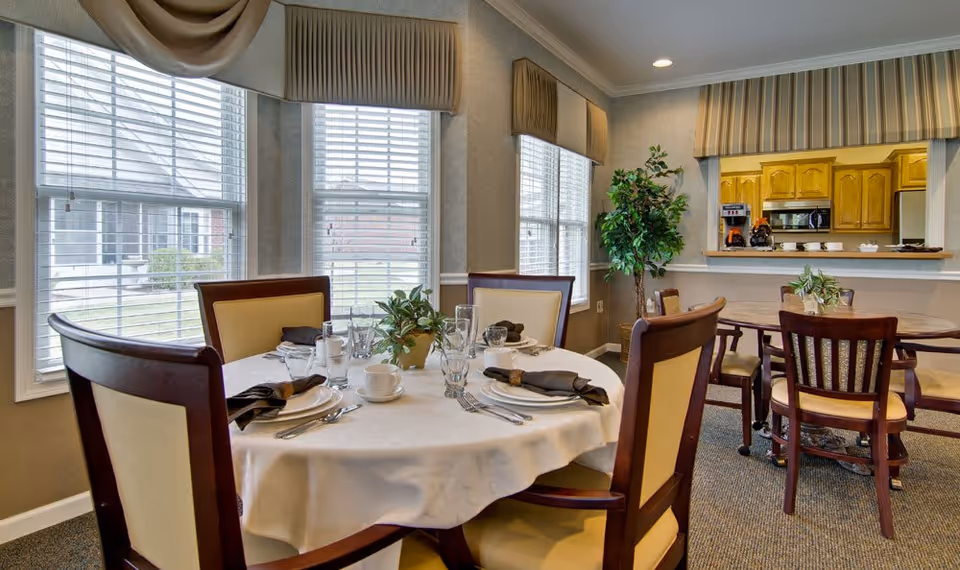 A dining area in a senior living facility with round tables covered with white tablecloths, set with plates, cups, glasses, and silverware. The room has large windows with blinds and valances, beige walls, and a view into a kitchen with wooden cabinets. There are several wooden chairs with cream-colored cushions around the tables and a potted plant in the corner.