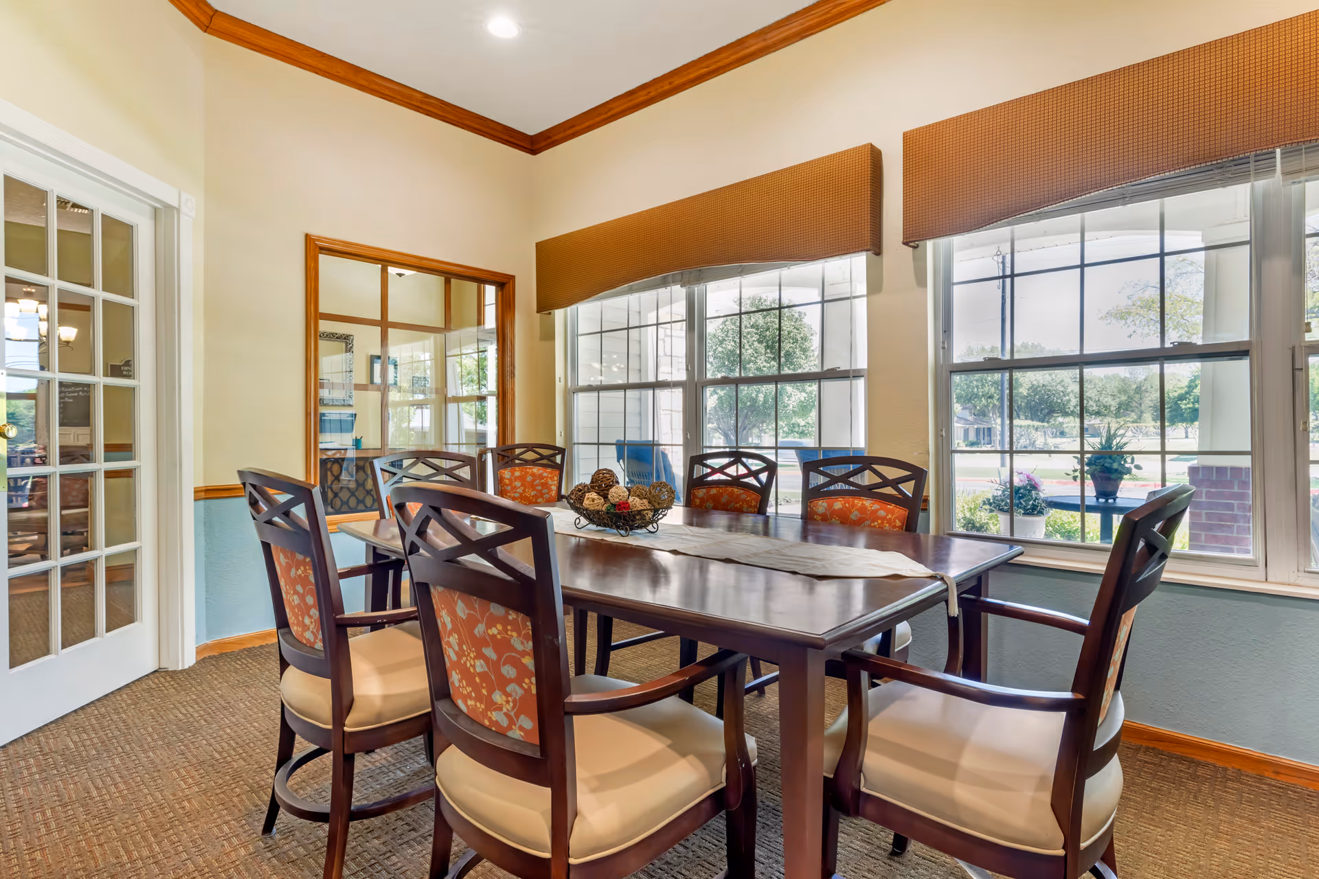 Sunlit dining room with a wooden table and upholstered chairs beside large windows overlooking landscaping.