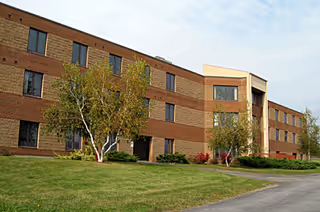 Exterior view of a three-story brick building with multiple windows, surrounded by green grass, small trees, and shrubs under a partly cloudy sky.