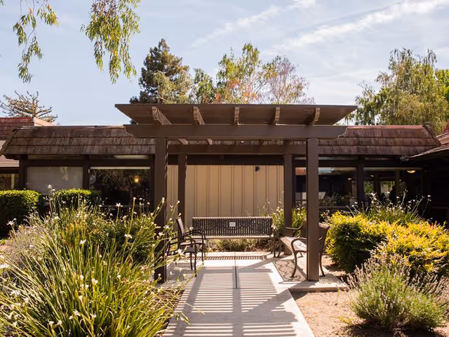 Outdoor garden area at Eskaton Care Center Greenhaven featuring a wooden pergola with benches underneath, surrounded by various green shrubs and plants, with a building in the background under a partly cloudy sky.