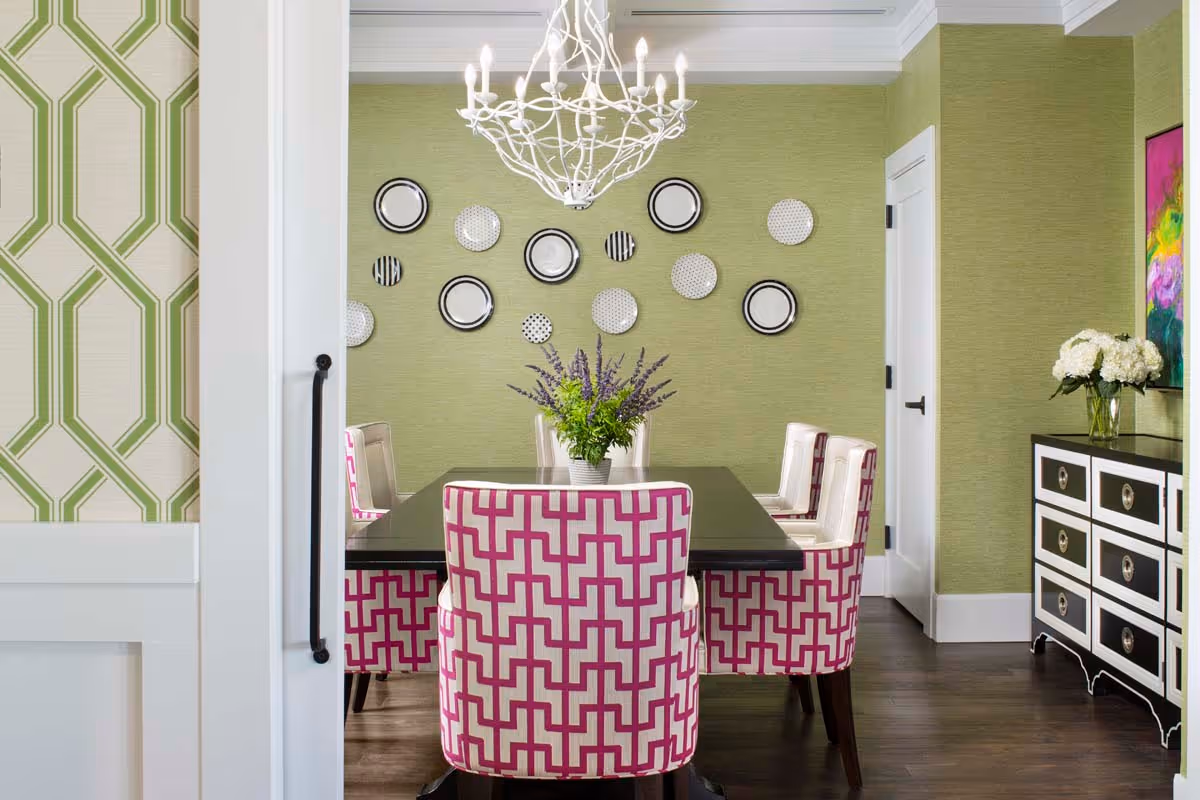 A stylish dining room with a black rectangular table surrounded by six chairs, four with white upholstery and two with a pink and white geometric pattern. The walls are painted light green with decorative plates arranged in a pattern on one wall. A white chandelier hangs above the table, and a black and white dresser with a vase of white flowers is on the right side. The floor is dark wood, and there is a colorful abstract painting on the right wall.