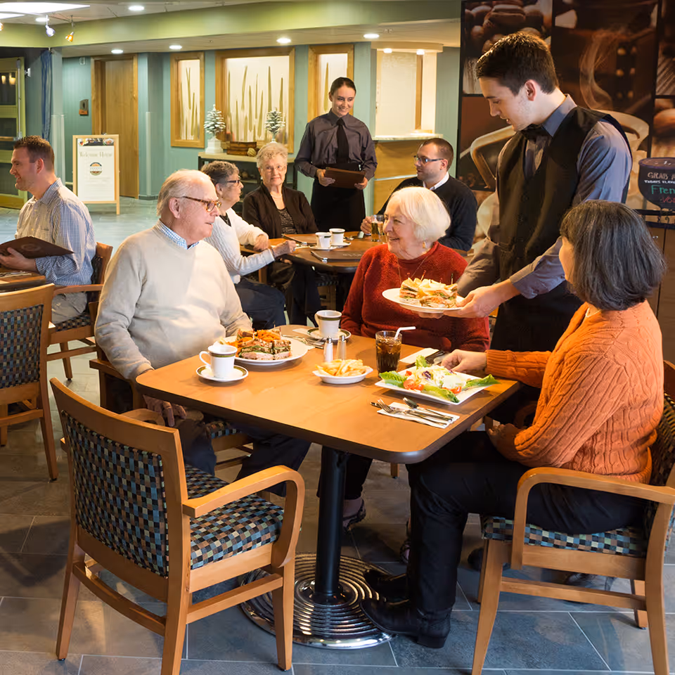 A group of elderly people sitting at tables in a dining area of a nursing home, being served food by waitstaff. The room has wooden chairs with patterned cushions, and the atmosphere is warm and social.