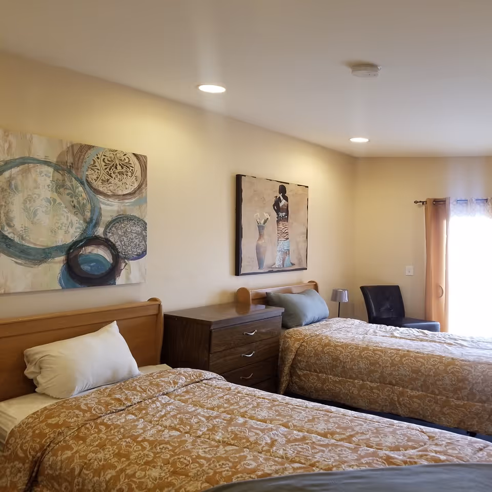 A bedroom with two single beds covered in patterned beige bedspreads. Between the beds is a wooden dresser with three drawers. Above each bed hangs a piece of artwork, one featuring abstract circular designs and the other depicting a figure with a vase. A black chair and a window with beige curtains are visible in the background. The room is softly lit by recessed ceiling lights.