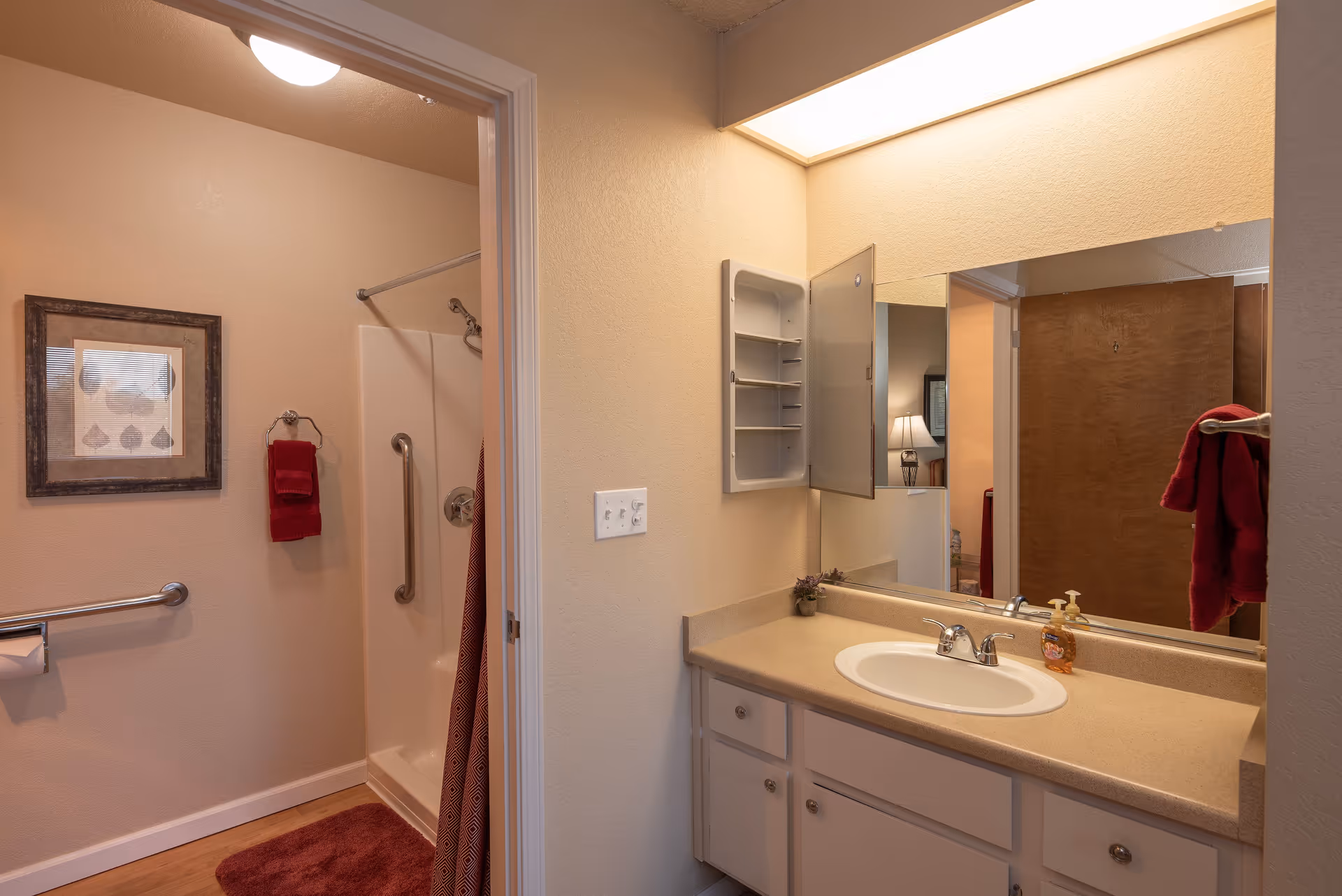 Interior view of a senior living facility bathroom with a sink and countertop on the right, a large mirror above the sink, and an open medicine cabinet. To the left is a shower area with grab bars, a red towel hanging on the wall, a framed picture, and a red bath mat on the floor.