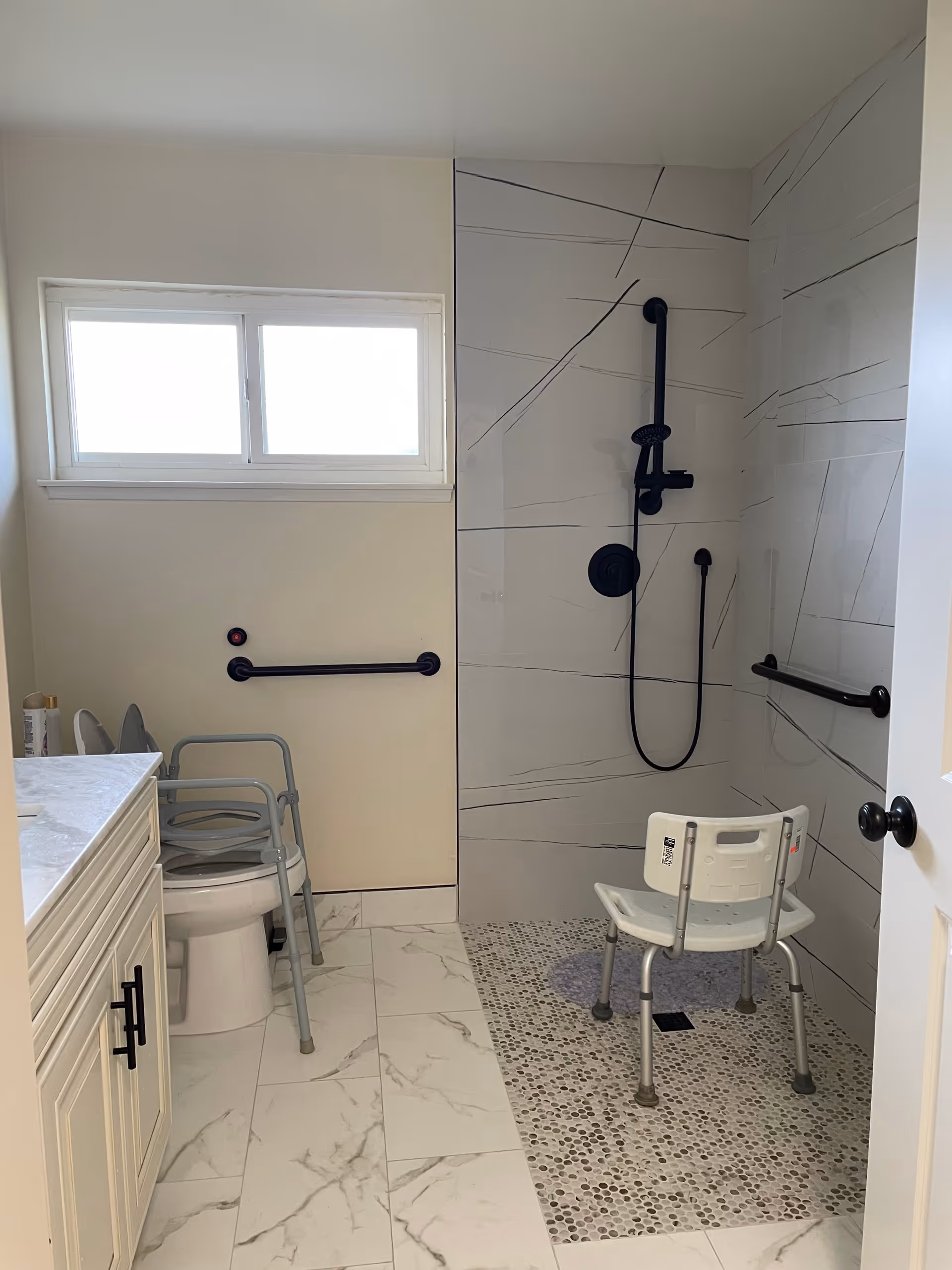 Accessible bathroom with a roll-in shower, shower chair, black grab bars, toilet with support frame, and marble-look tile floor.