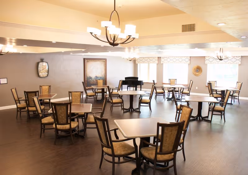 A spacious dining room with multiple square tables and cushioned chairs arranged neatly on a dark wooden floor. The room has beige walls, large windows letting in natural light, decorative wall art, and several modern chandeliers hanging from the ceiling. A black piano is positioned near the windows.