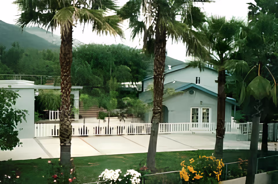 Light-colored building front with palm trees, a white picket fence, and landscaped lawn and flowers.