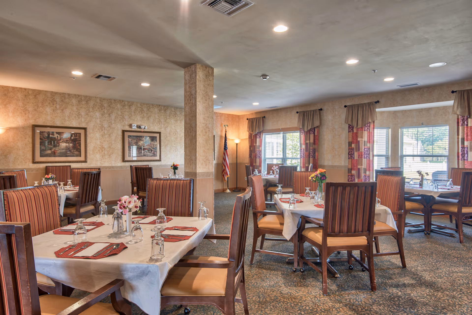 A dining room with several tables covered with white tablecloths, each set with glasses, napkins, and floral centerpieces. The room has striped upholstered chairs, patterned curtains on the windows, framed artwork on the walls, and an American flag in the corner.