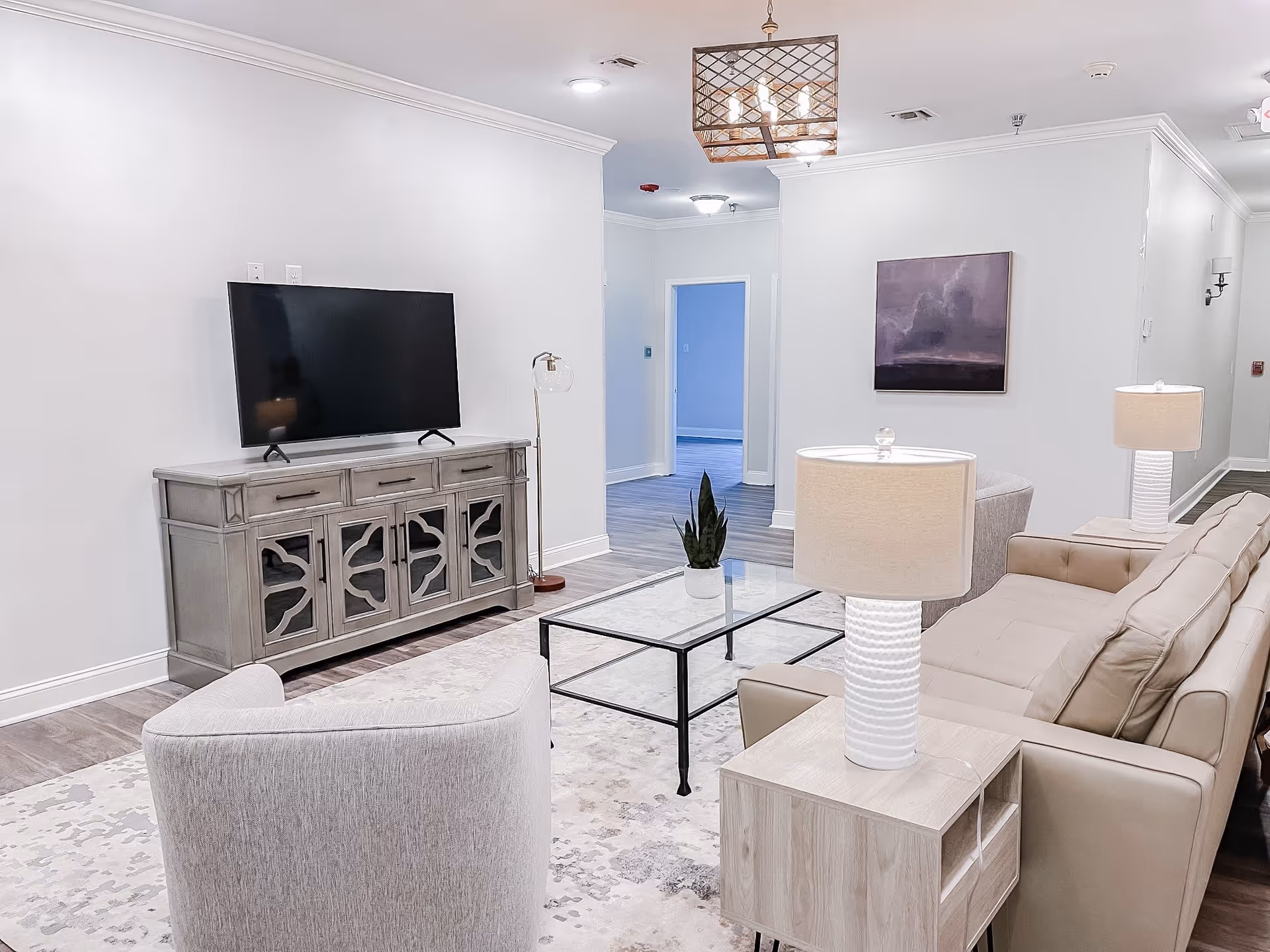 A modern living room with a beige sofa, a light gray armchair, a glass coffee table with a small plant, two table lamps with white textured bases, a flat-screen TV on a gray wooden cabinet, and a purple abstract painting on the wall. The room has light-colored walls and wood flooring.