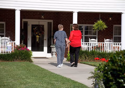 Two women walking hand in hand toward the front entrance of a brick retirement center with a porch and rocking chairs.