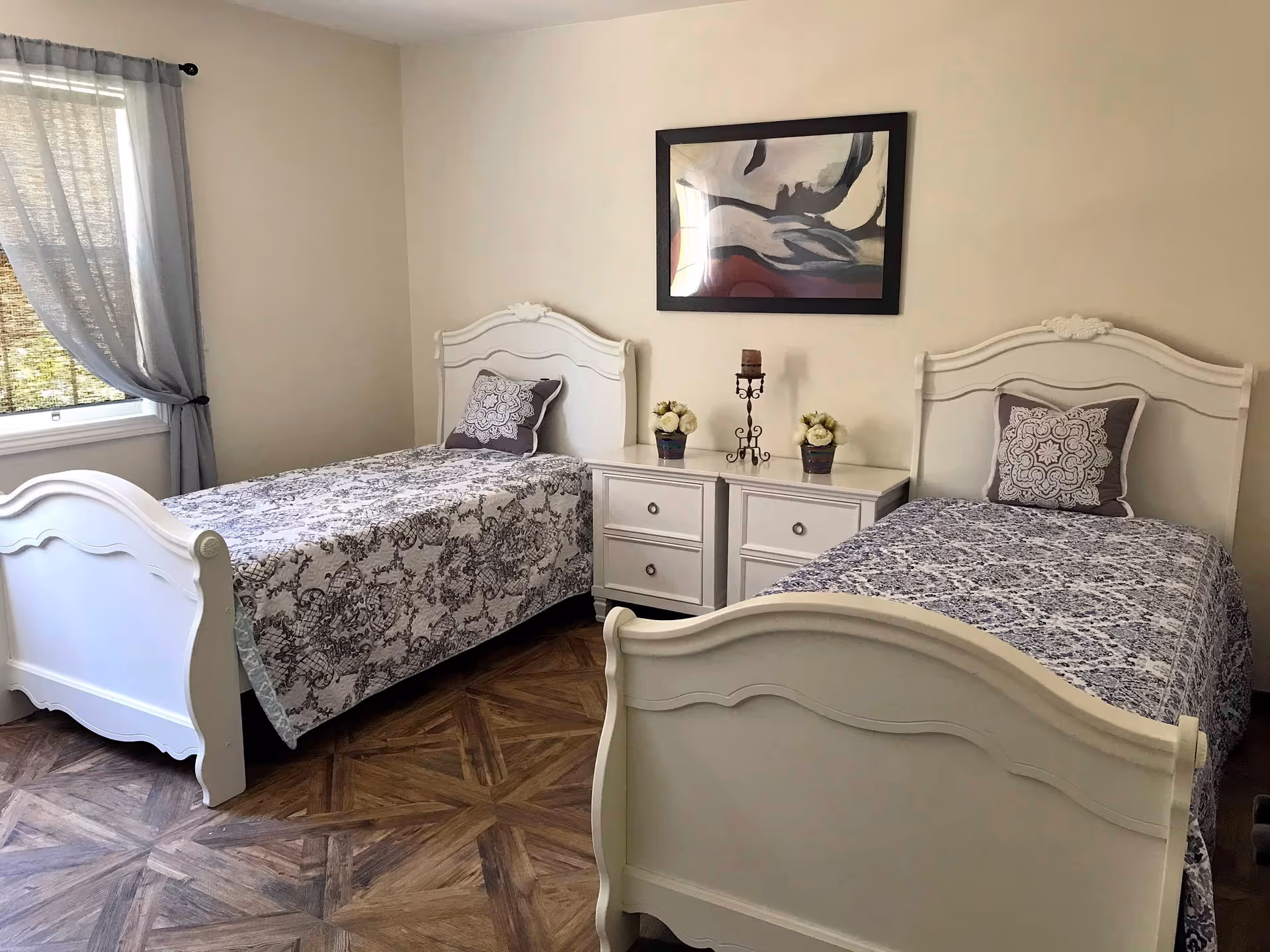 A bedroom with two white twin beds featuring patterned bedspreads and matching decorative pillows. Between the beds is a white nightstand with two drawers, decorated with two small flower pots and a candle holder. A large abstract painting hangs on the beige wall above the nightstand. A window with sheer gray curtains is on the left side of the room, and the floor has a wood-patterned design.